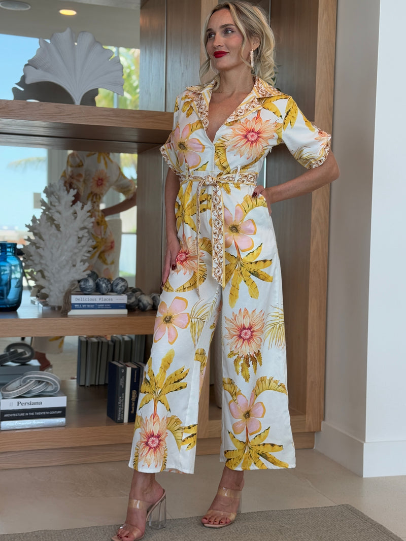 Woman in belted tropical floral jumpsuit with soft yellow and blush print.